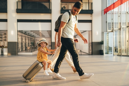 A father pulling a suitcase with his young child riding on it while traveling through an airport, illustrating out-of-state holiday travel with children for Virginia parents.